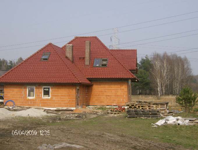 Dom w trakcie budowy z czerwoną dachówką, widoczne okna dachowe i komin, ściany z cegły dziurawki, na pierwszym planie palety i gruz, w tle las i słupy wysokiego napięcia.